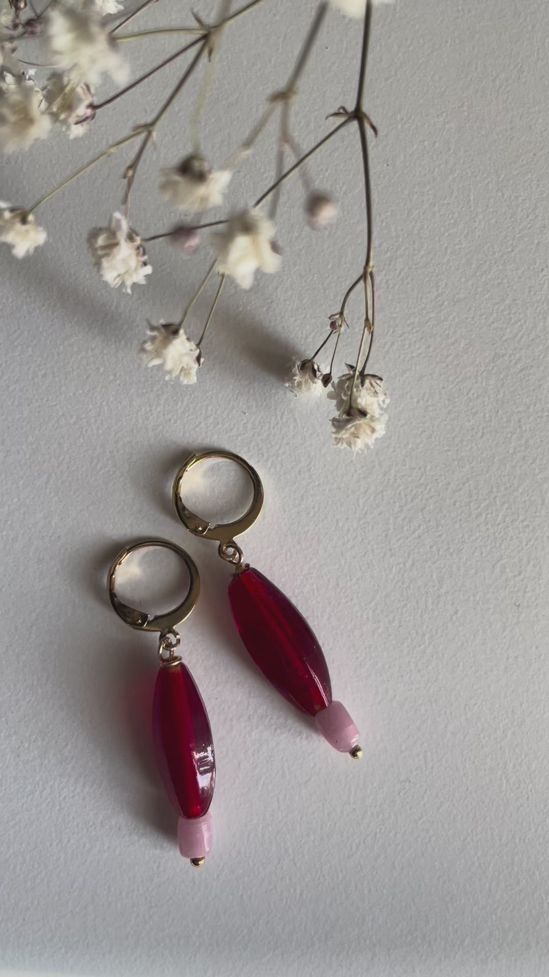 Red Earrings, A pair of gold plated huggie hoops with red elongated glass bead charms with small pastel pink beads shown on a white surface with natural sunlight.