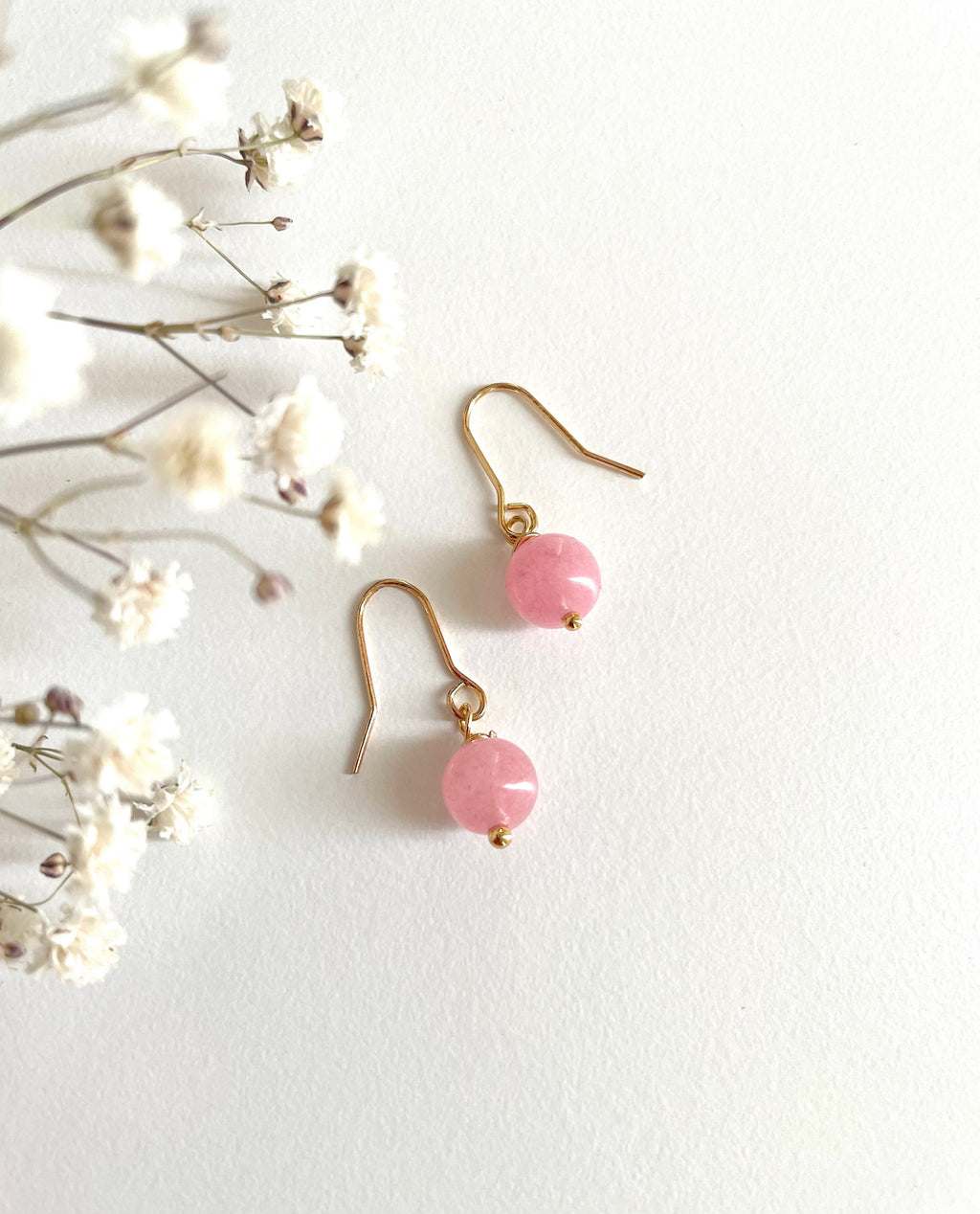 A pair of gold plated drop earrings with round pink natural rose quartz gemstone beads and gold dainty hooks, displayed on a textured white background next to delicate white dried flowers.