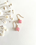 A pair of gold plated drop earrings with round pink natural rose quartz gemstone beads and gold dainty hooks, displayed on a textured white background next to delicate white dried flowers.