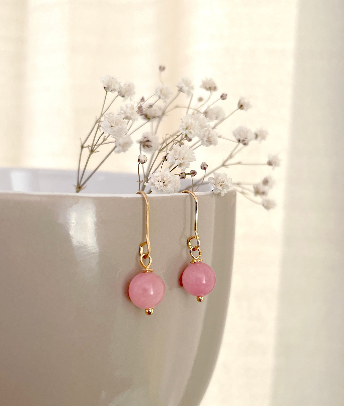 A pair of gold plated drop earrings with round light pink beads made of real rose quartz gemstone hanging from the rim of a light beige ceramic bowl, with small white dried flowers placed above.