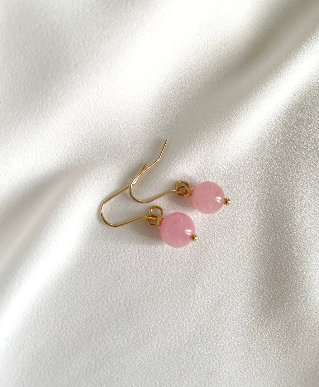 A pair of light pink earrings with gold plated dangle hooks with smooth, round pastel pink beads of natural rose quartz gemstone, displayed on a softly draped white fabric background.