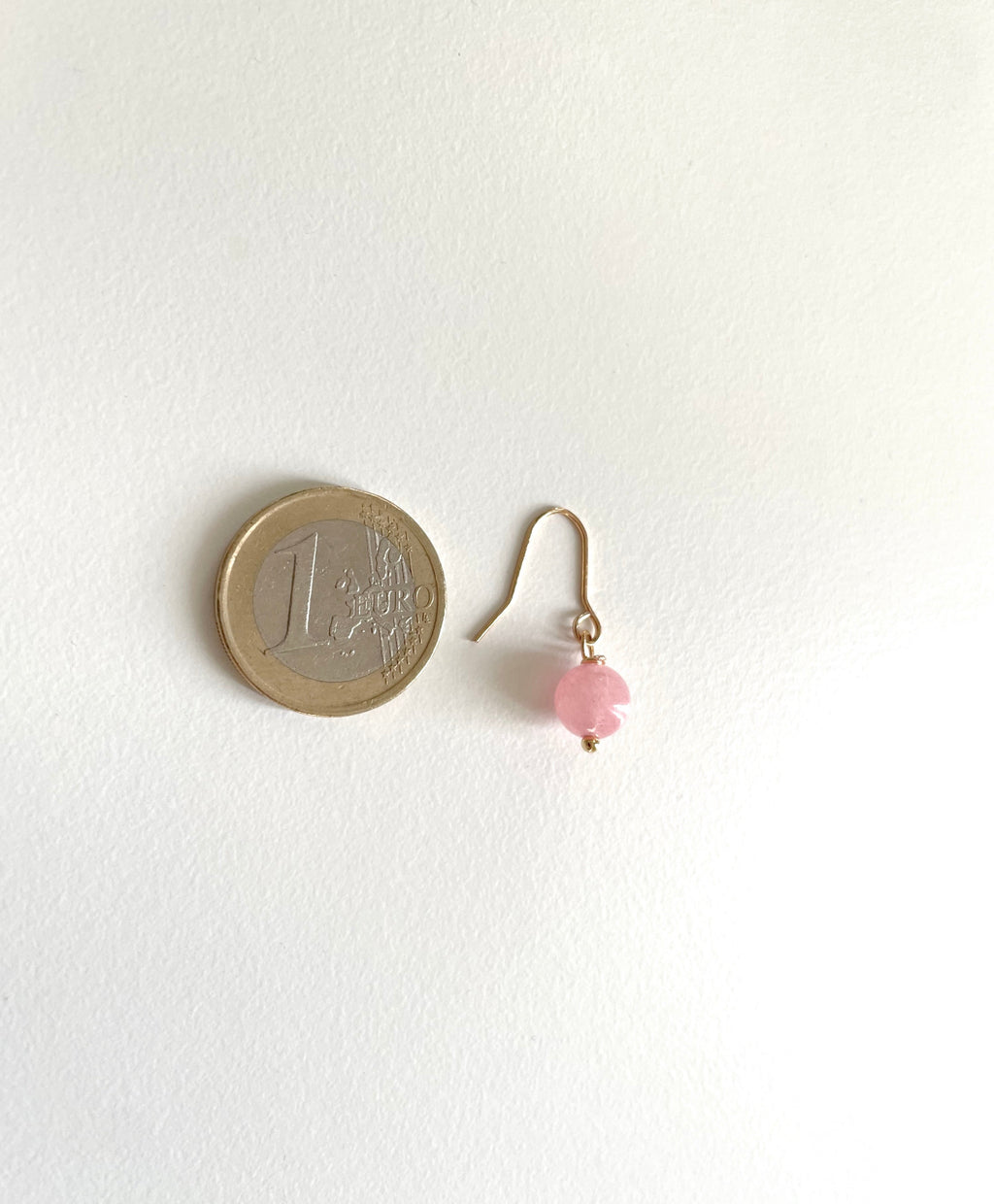 A single small drop earring with a gold plated fishhook wire and a tiny pastel pink bead, shown next to a 1 euro coin for scale on a plain white background.