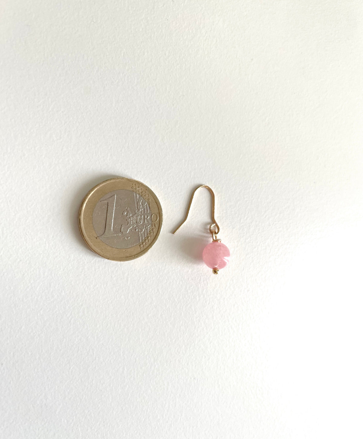 A single small drop earring with a gold plated fishhook wire and a tiny pastel pink bead, shown next to a 1 euro coin for scale on a plain white background.
